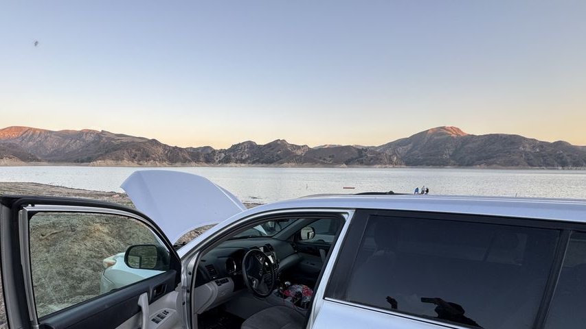 An SUV with its hood up and driver door open on the shore of a lake at sunset, mountains behind