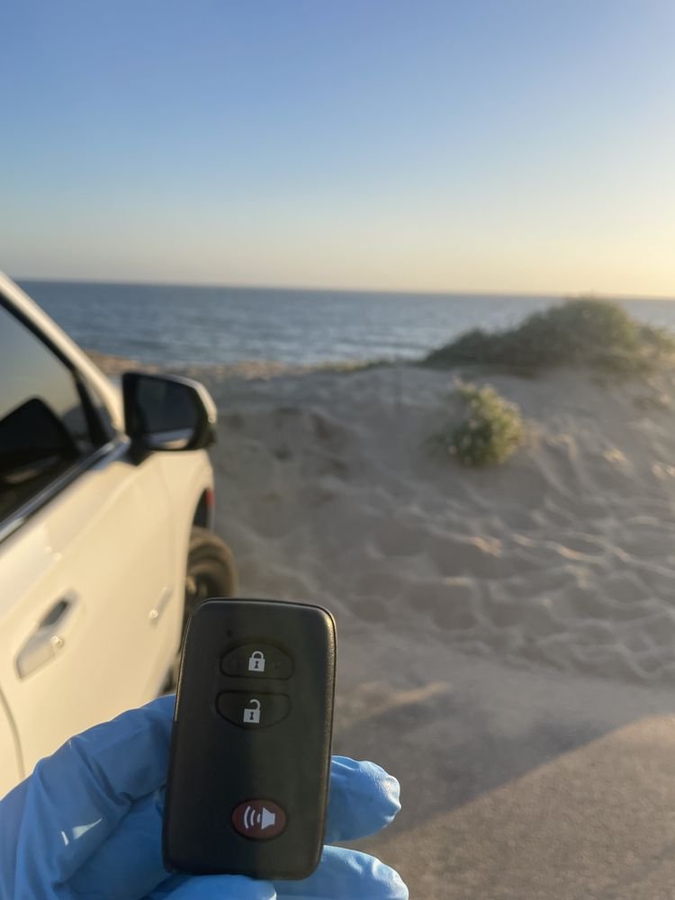 A smart key in a gloved hand beside a white car, the ocean and sand dunes behind